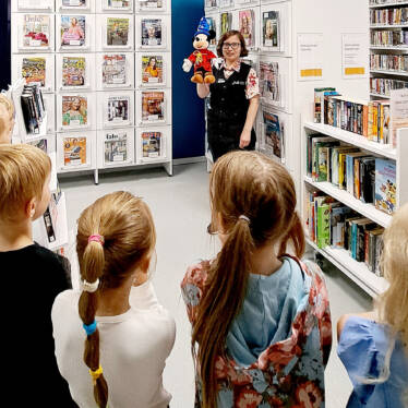 Förskolelever på biblioteksbesök, bibliotekarien har ett Musse Pigg -kramdjur i handen.