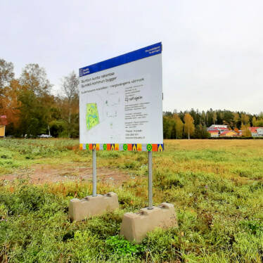 En byggarbetsplatsskylt i förgrunden framför en åker i träda, bakom åkern syns villor.