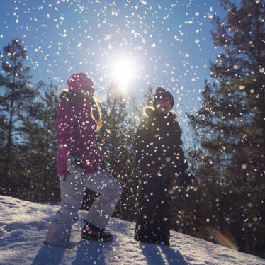 Två barn står på en snöig sluttning med solen i bakgrunden, i förgrunden yr snöflingor