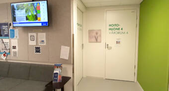 Odotusaulassa infonäyttö ja terveydenhoitajan ovi, jossa lukee Hoitohuone 4.