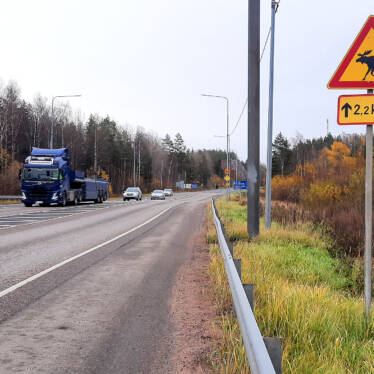 På bilden stamväg 51, bilar och en långtradare på vägen, varning för älgar på vägrenen.