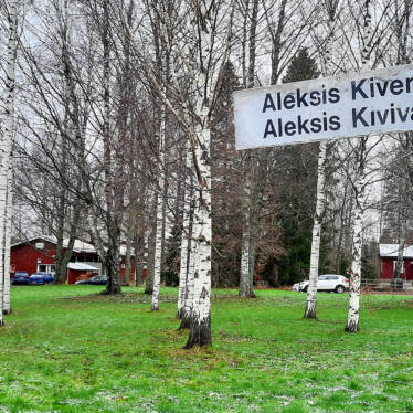 På bilden finns en björkpark och bakom den syns daghemmet Jukola till vänster och Fanjunkars torp till höger. Framför torpets gård ligger en parkeringsplats och i förgrunden på bilden finns en gatunamnsskylt där det står Aleksis Kivivägen.